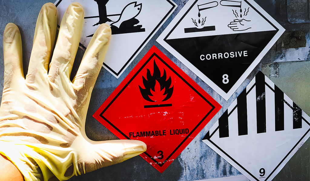 A hand in a glove placed against a barrel which features hazardous waste stickers
