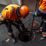 Two workers in high vis clothing placing two hoses down a manhole