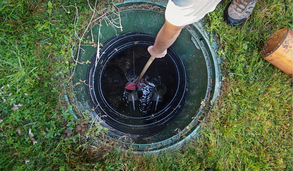 Person inspecting an open septic system access point, using a tool to check wastewater levels and components inside the tank during maintenance or inspection.